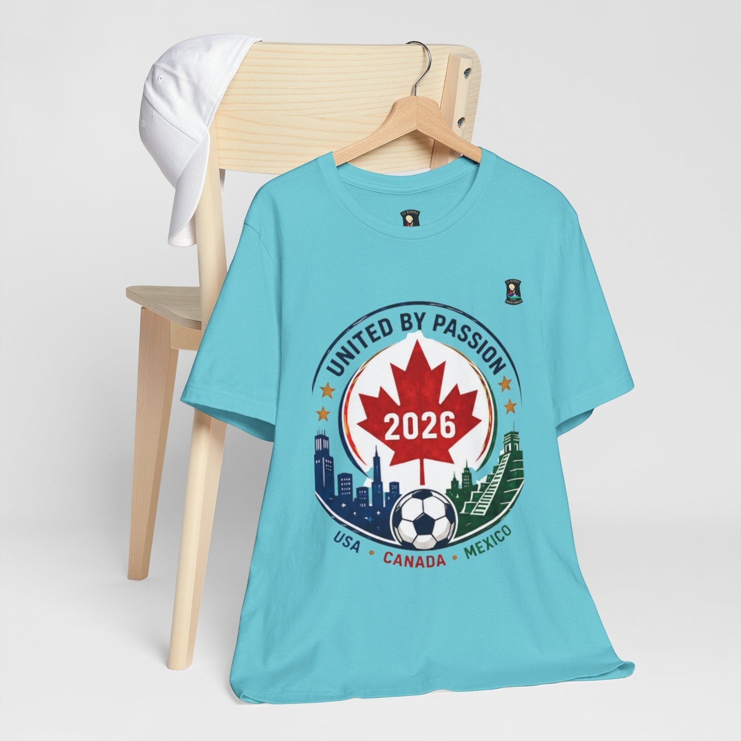 CJ Riders Foundation Store light blue 100% cotton t-shirt with a circular graphic celebrating the 2026 World Cup, featuring a Canadian maple leaf, soccer ball, and skylines of USA, Canada, and Mexico.