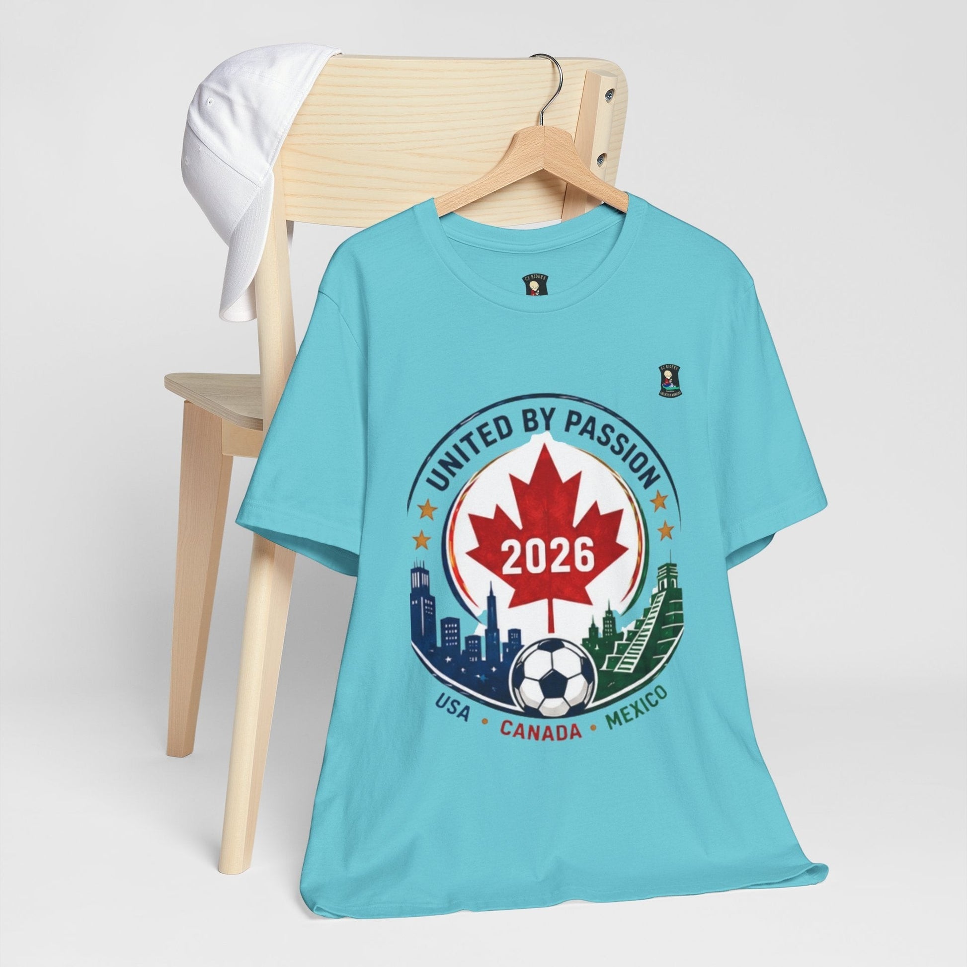 CJ Riders Foundation Store light blue 100% cotton t-shirt with a circular graphic celebrating the 2026 World Cup, featuring a Canadian maple leaf, soccer ball, and skylines of USA, Canada, and Mexico.