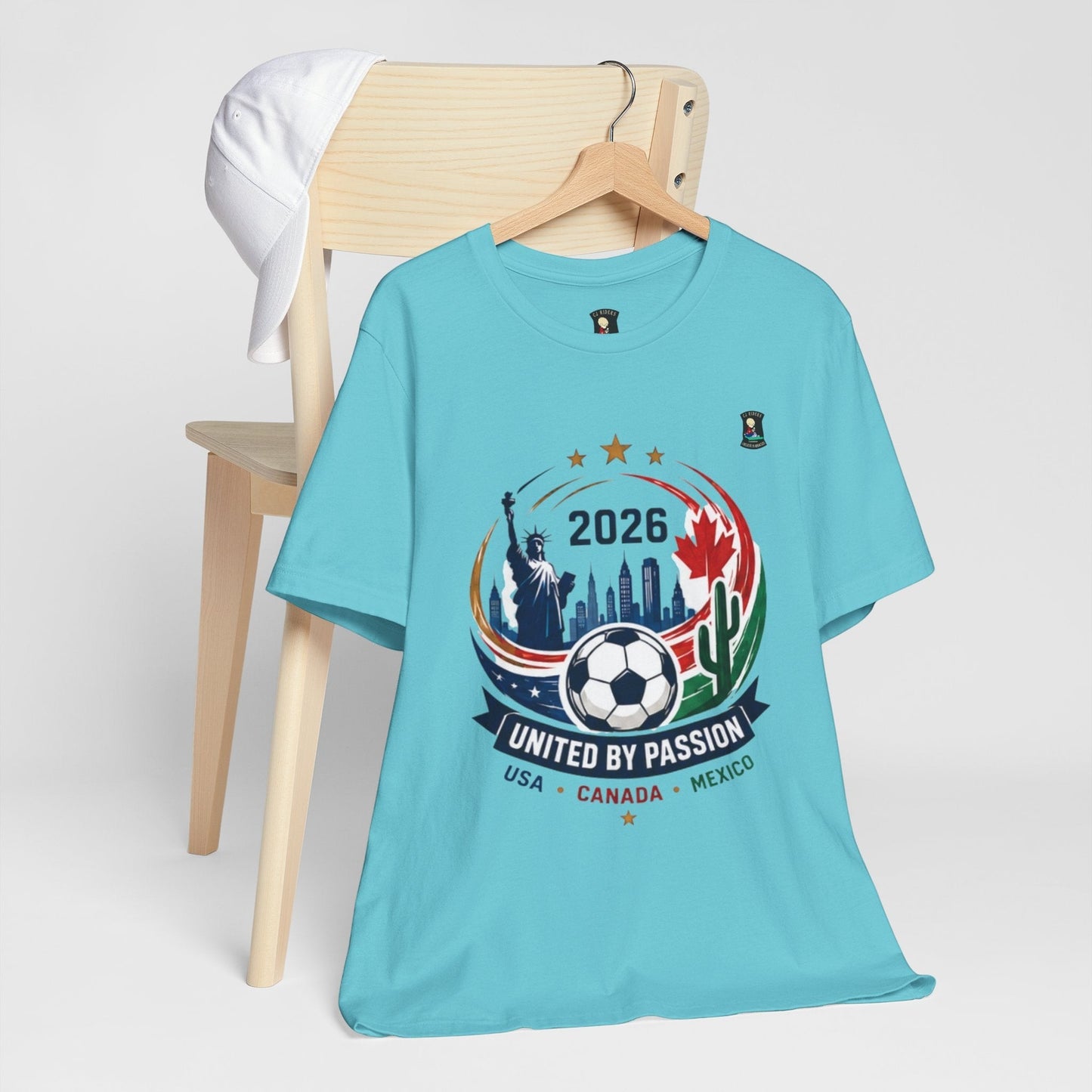 CJ Riders Foundation Store light blue 100% cotton t-shirt with 2026 soccer graphic featuring Statue of Liberty, skyline, soccer ball, and flags.