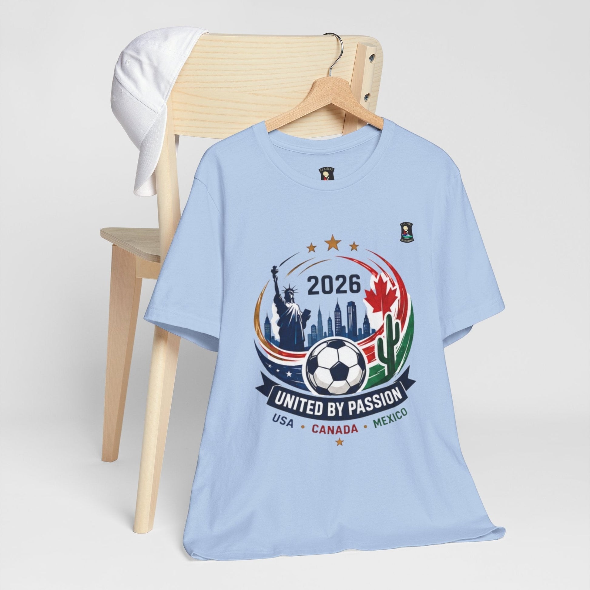 CJ Riders Foundation Store light blue 100% cotton t-shirt with 2026 soccer graphic featuring Statue of Liberty, Canadian maple leaf, Mexican cactus, and soccer ball.