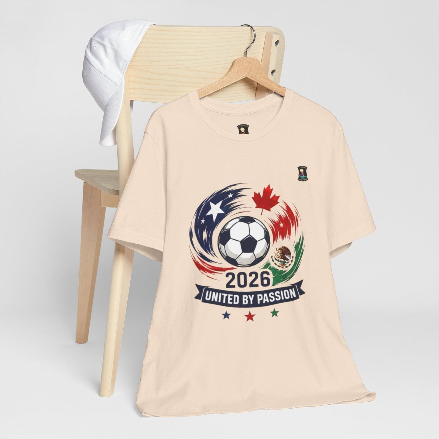 CJ Riders Foundation Store cream 100% cotton t-shirt with 2026 soccer graphic featuring USA, Canada, Mexico flags and "United by Passion" text.