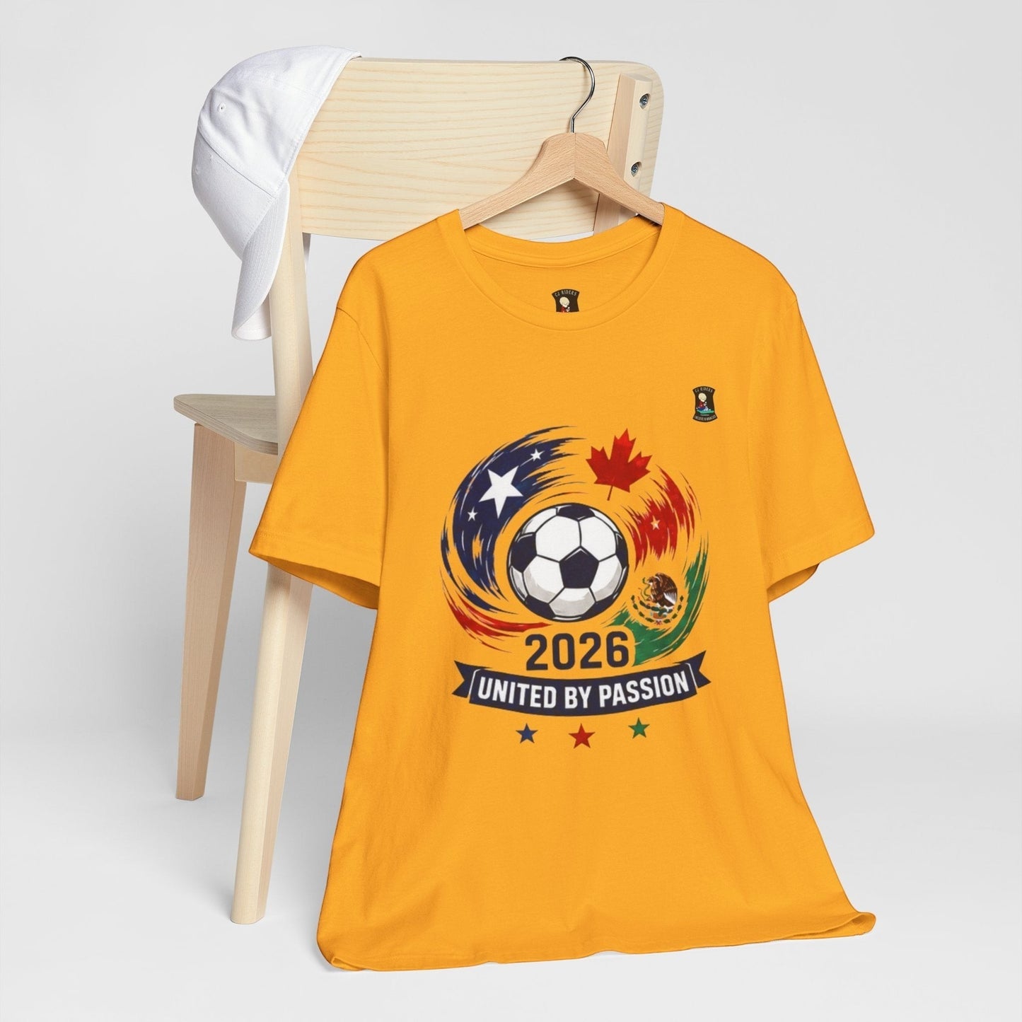 CJ Riders Foundation Store yellow 100% cotton t-shirt with 2026 soccer graphic, featuring a ball, USA, Canada, Mexico flags, and "United by Passion" text.
