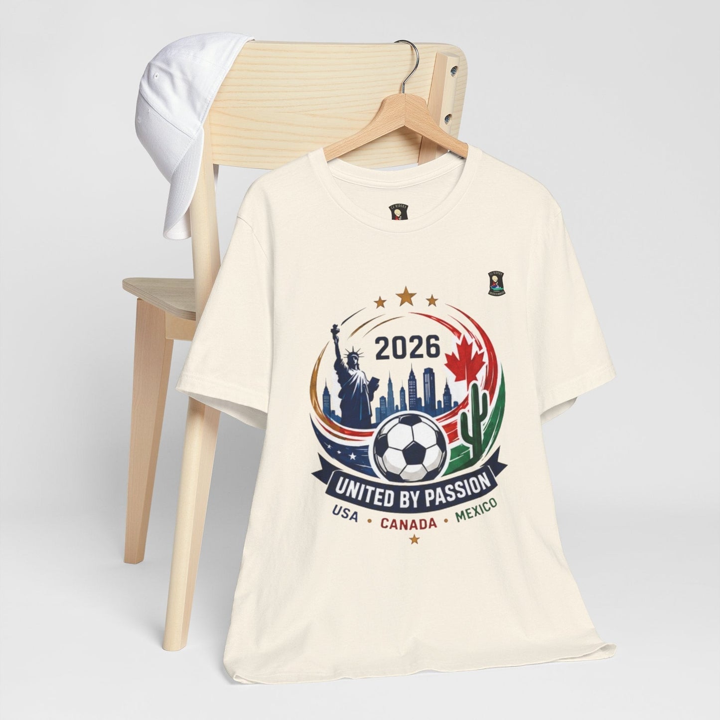 CJ Riders Foundation Store cream 100% cotton t-shirt with 2026 soccer graphic featuring Statue of Liberty, cityscape, soccer ball, and USA, Canada, Mexico flags.