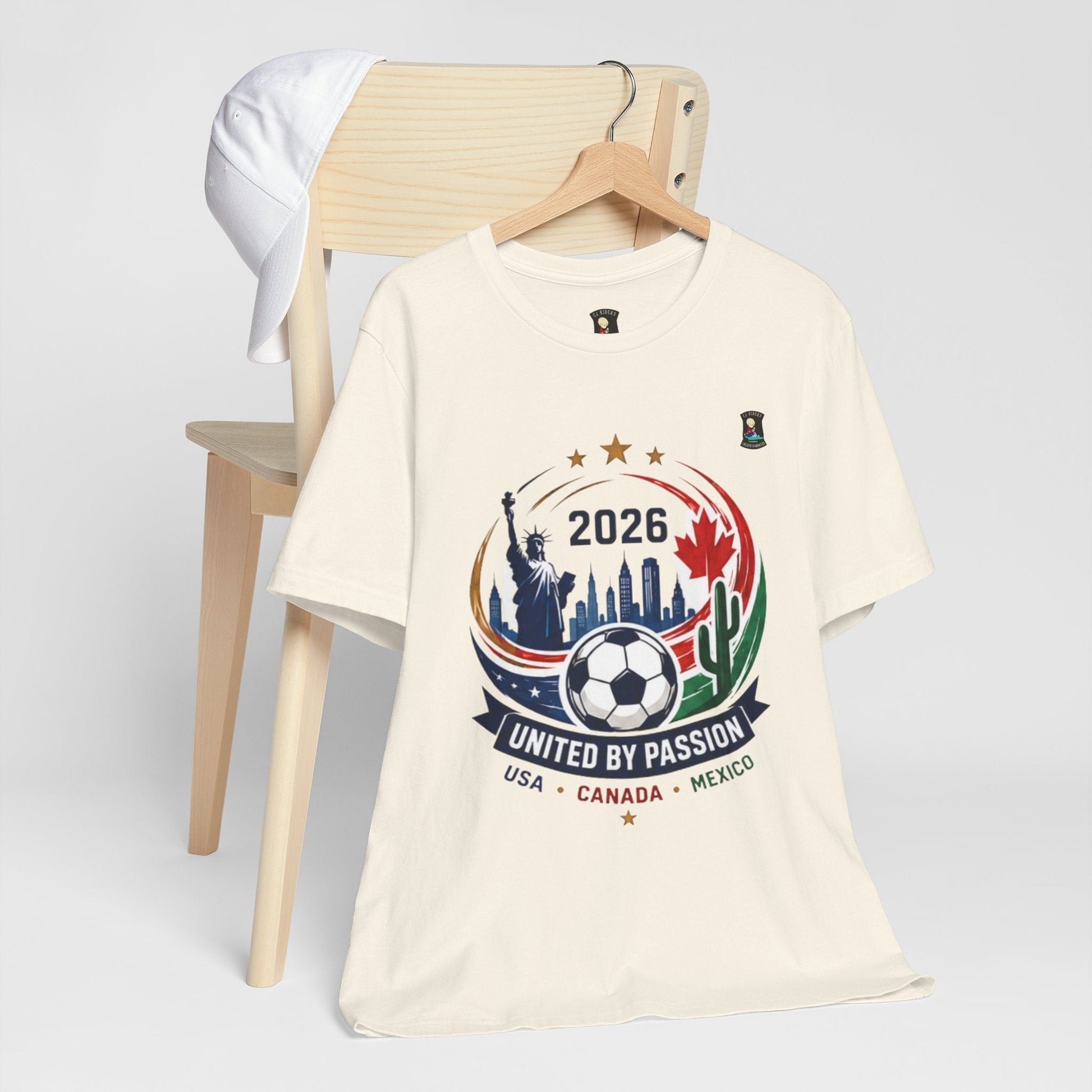 CJ Riders Foundation Store cream 100% cotton t-shirt with 2026 soccer graphic featuring Statue of Liberty, cityscape, soccer ball, and USA, Canada, Mexico flags.