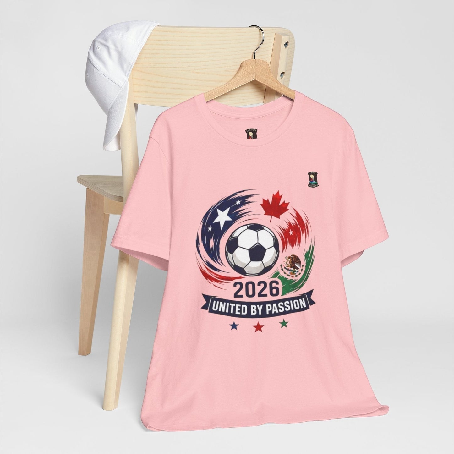 CJ Riders Foundation Store pink 100% cotton t-shirt with 2026 soccer graphic featuring a ball, USA, Canada, Mexico flags, and motion strokes.