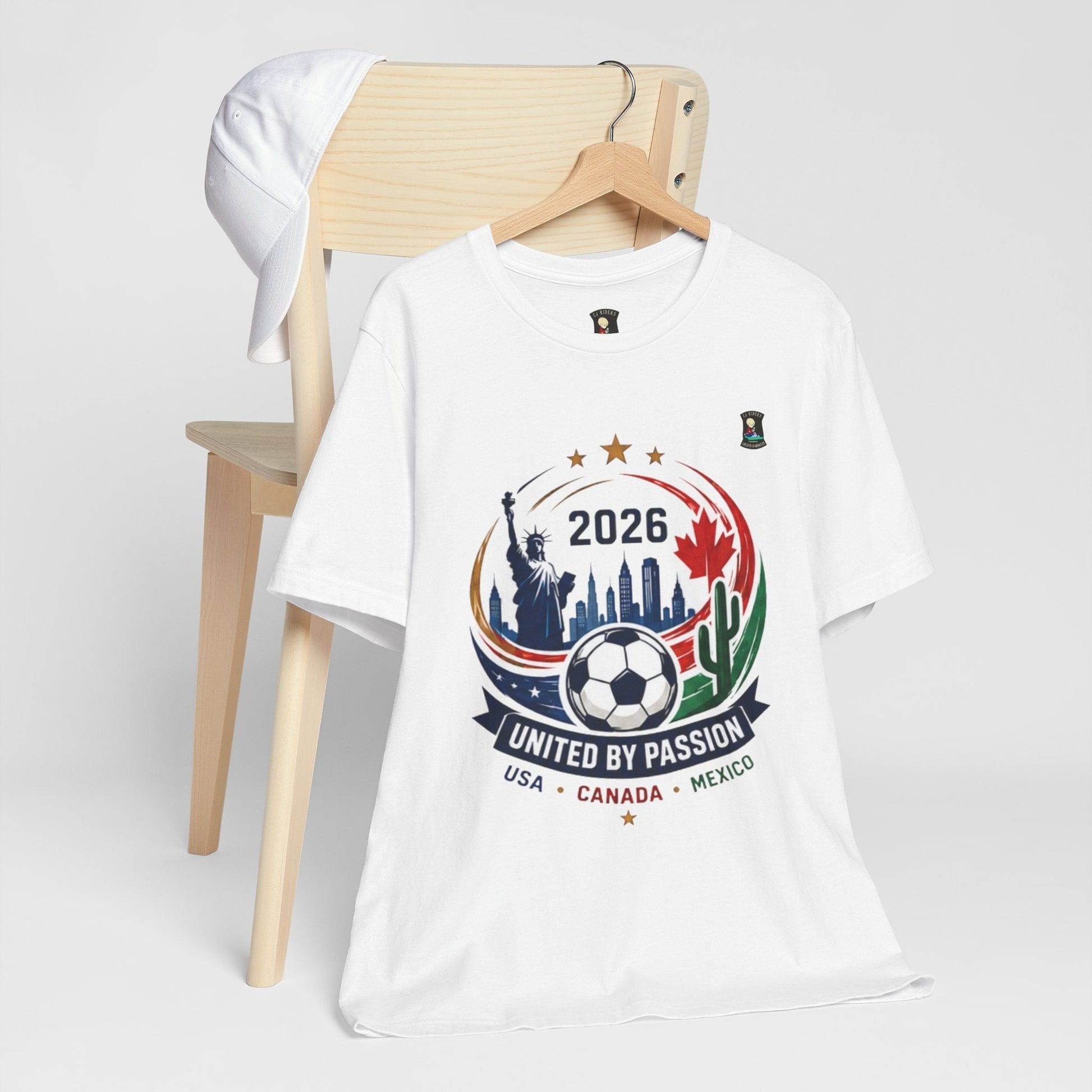 CJ Riders Foundation Store white 100% cotton t-shirt with 2026 soccer graphic featuring Statue of Liberty, cityscape, soccer ball, and USA, Canada, Mexico flags.