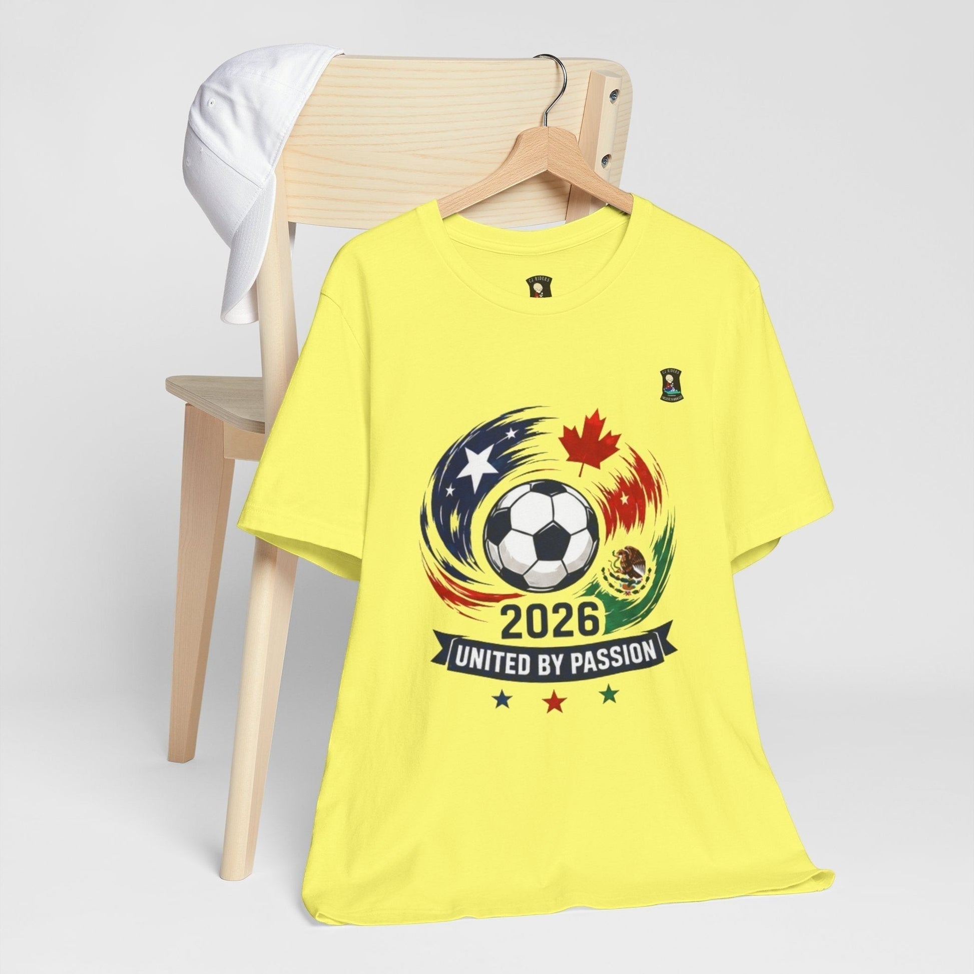 CJ Riders Foundation Store yellow 100% cotton t-shirt with 2026 soccer graphic featuring a ball, USA, Canada, Mexico flags, and "United by Passion" text.
