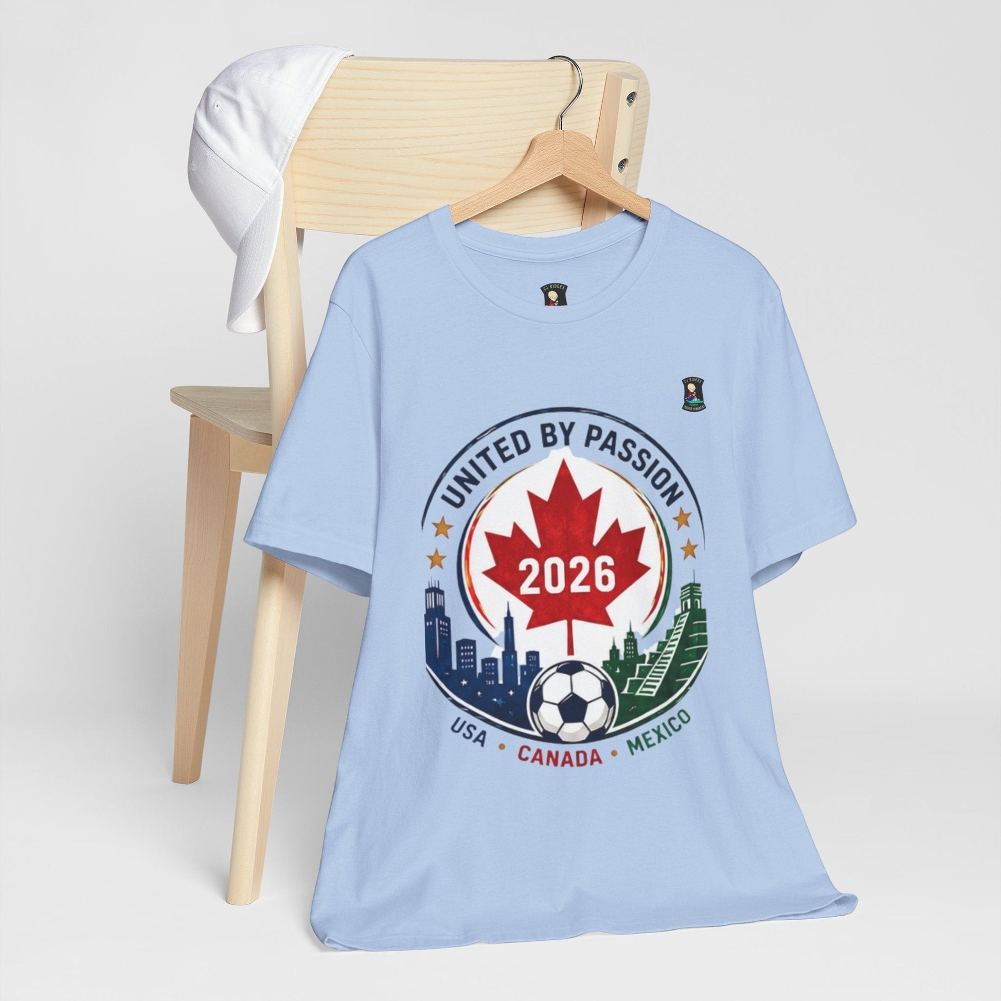 CJ Riders Foundation Store light blue 100% cotton t-shirt with 2026 soccer crest design featuring Canadian maple leaf, soccer ball, and city skylines.
