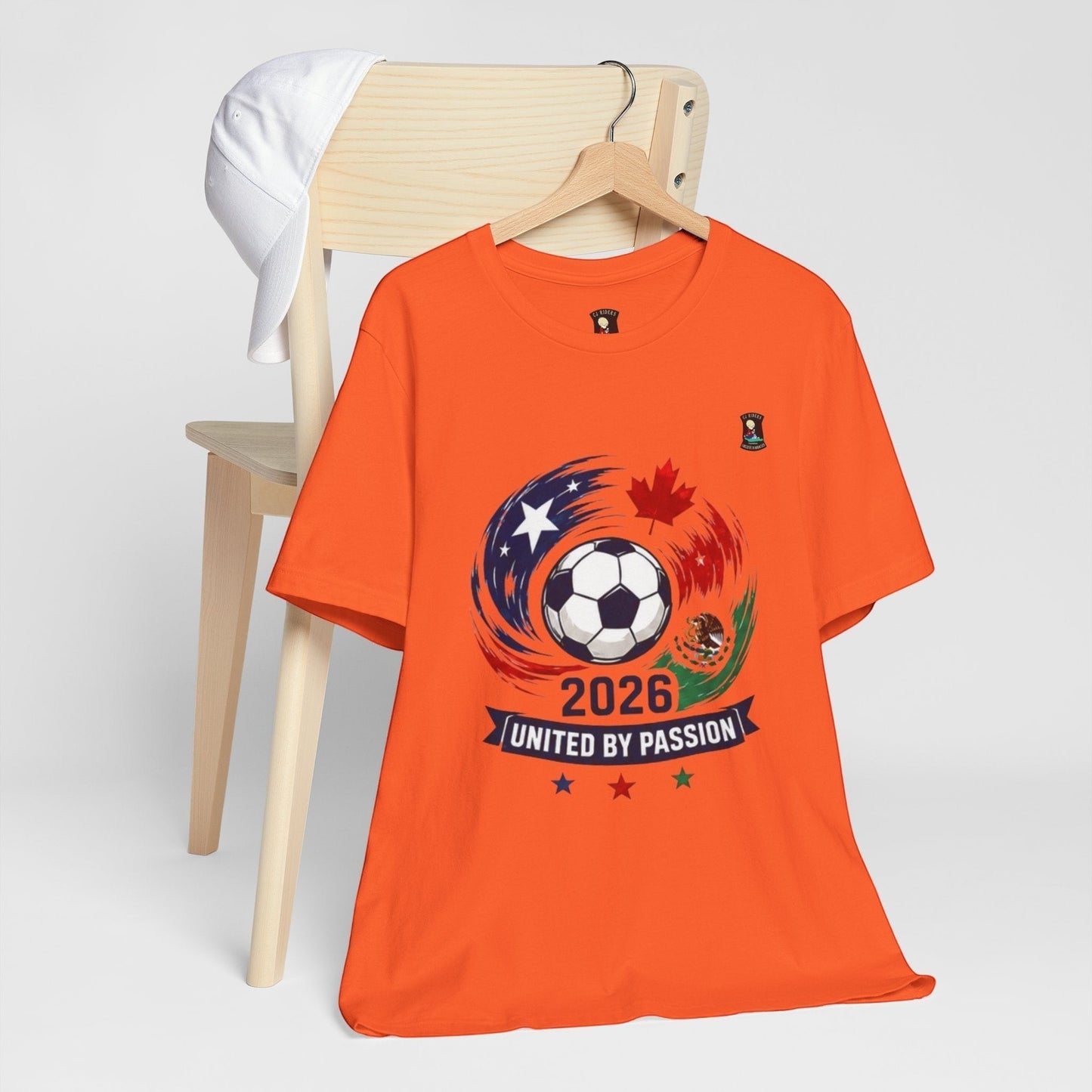 CJ Riders Foundation Store orange 100% cotton t-shirt with 2026 soccer graphic featuring a ball, USA, Canada, Mexico flags, and "United by Passion" text.