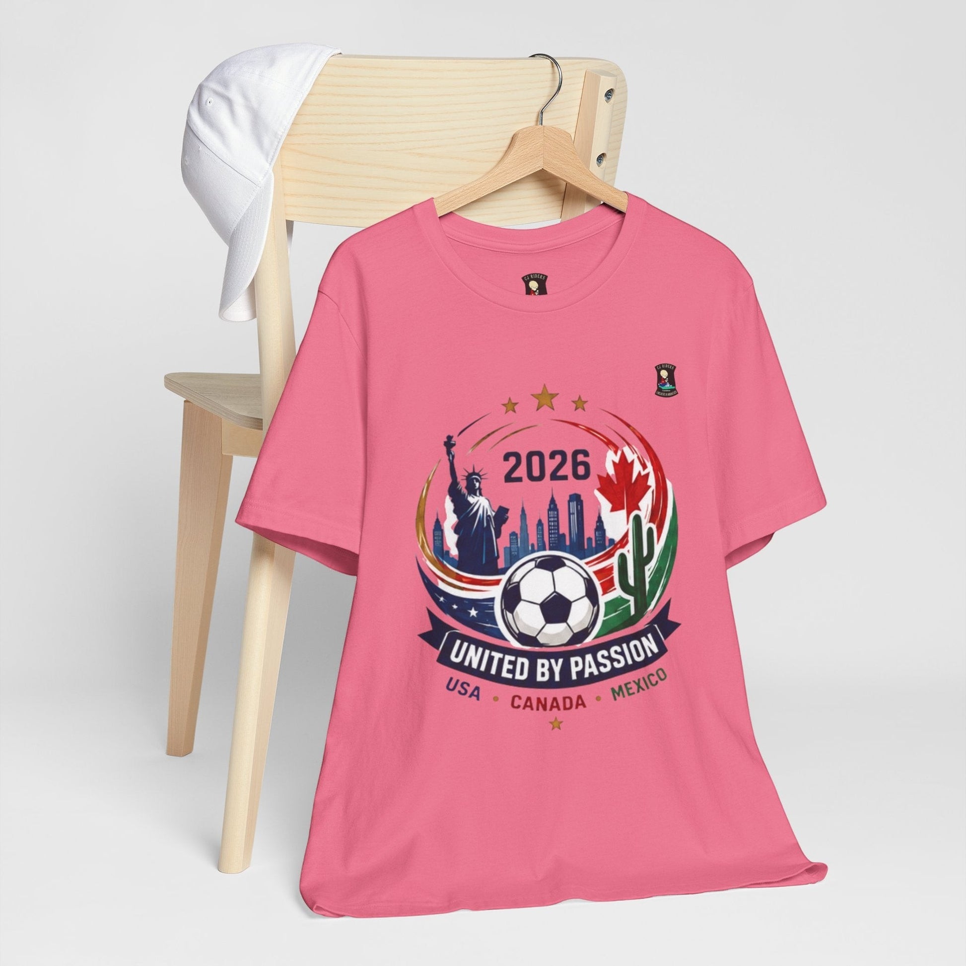 CJ Riders Foundation Store pink 100% cotton t-shirt with 2026 World Cup graphic featuring Statue of Liberty, Canadian maple leaf, and soccer ball.