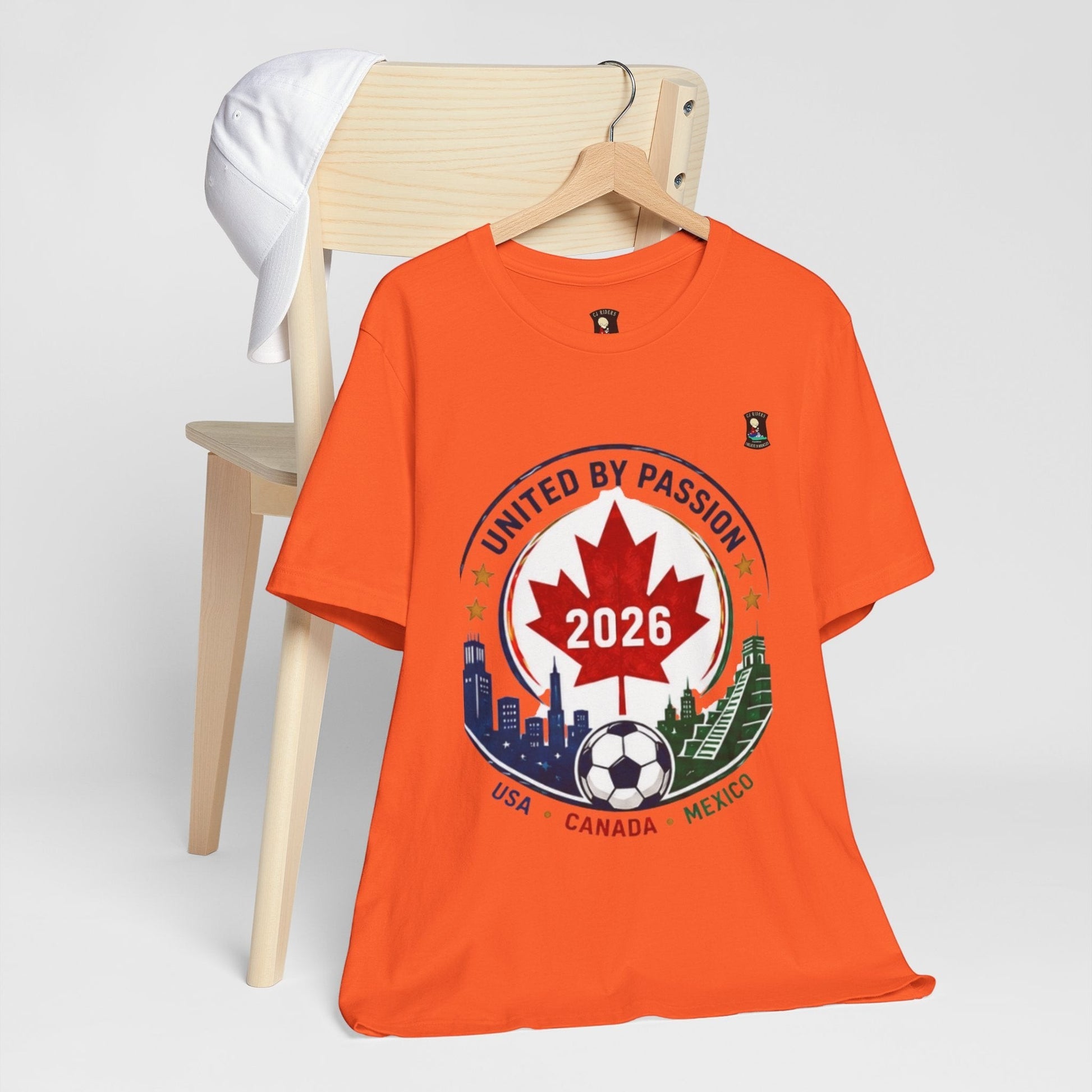 CJ Riders Foundation Store orange 100% cotton t-shirt with a 2026 soccer crest design featuring a maple leaf, soccer ball, and city skylines of USA, Canada, and Mexico.