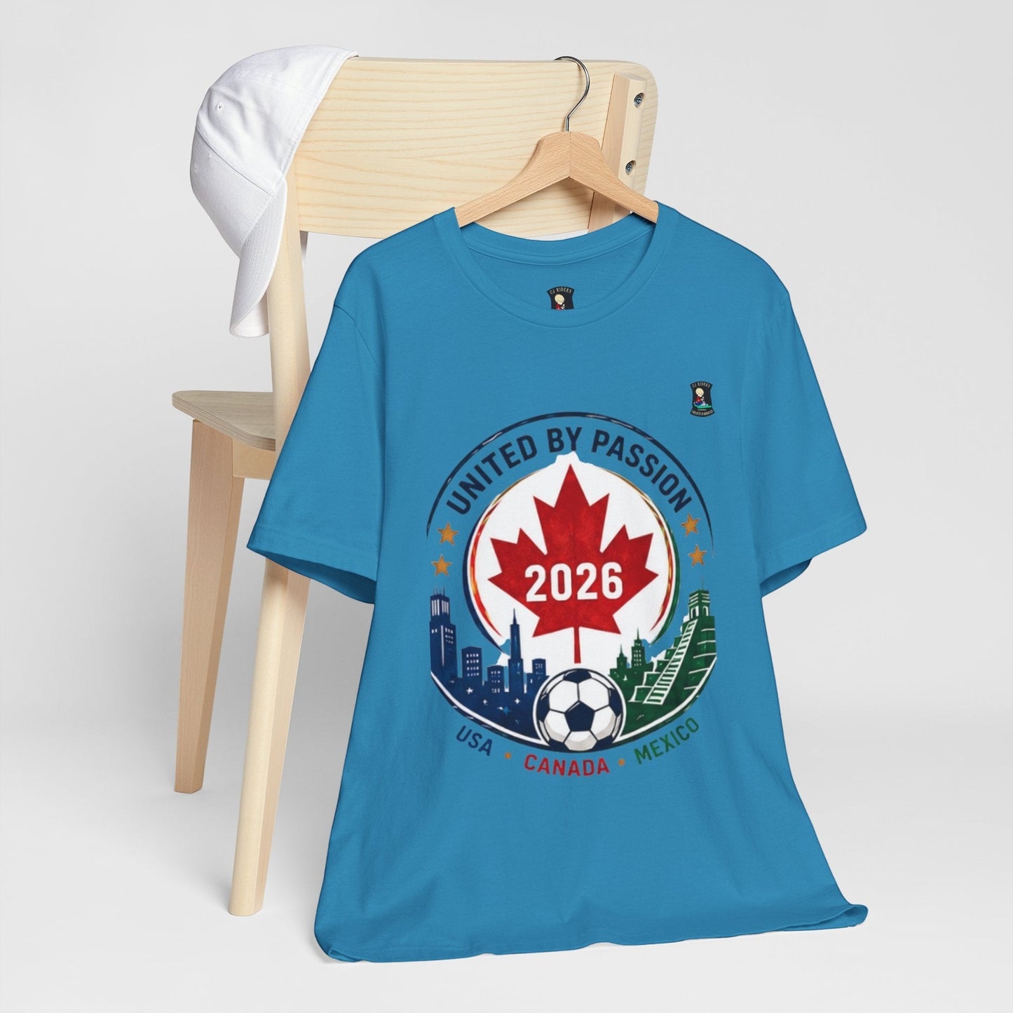 CJ Riders Foundation Store blue 100% cotton t-shirt with United by Passion 2026 soccer graphic featuring Canadian maple leaf, soccer ball, and city skylines of USA, Canada, Mexico.