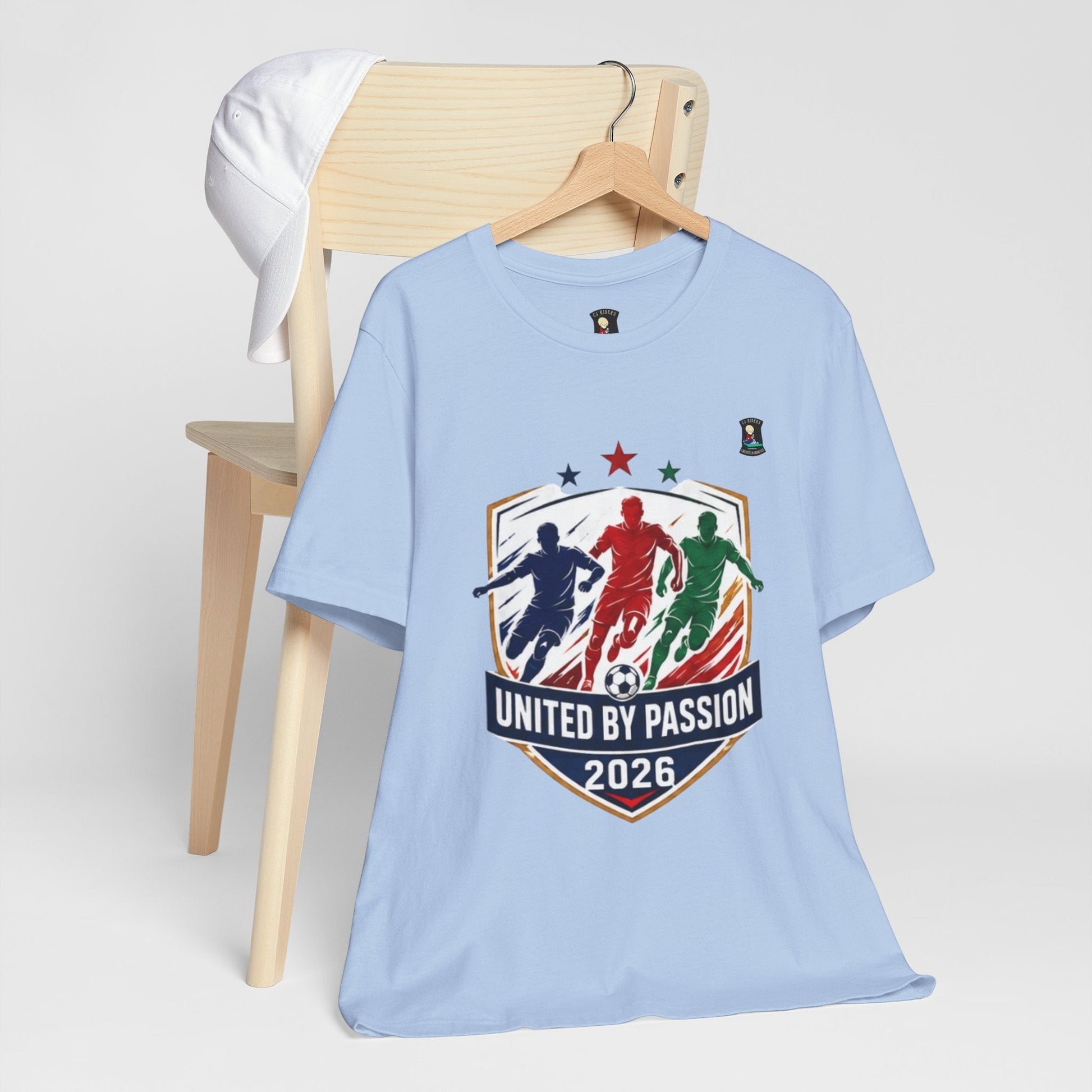 CJ Riders Foundation Store light blue 100% cotton t-shirt with "United by Passion 2026" graphic featuring three soccer players and a ball.