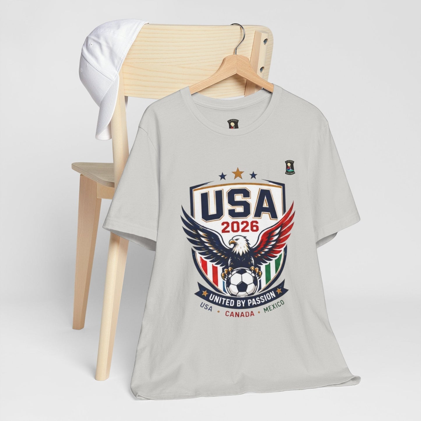 CJ Riders Foundation Store light grey 100% cotton t-shirt with USA 2026 eagle graphic and "United by Passion" text.