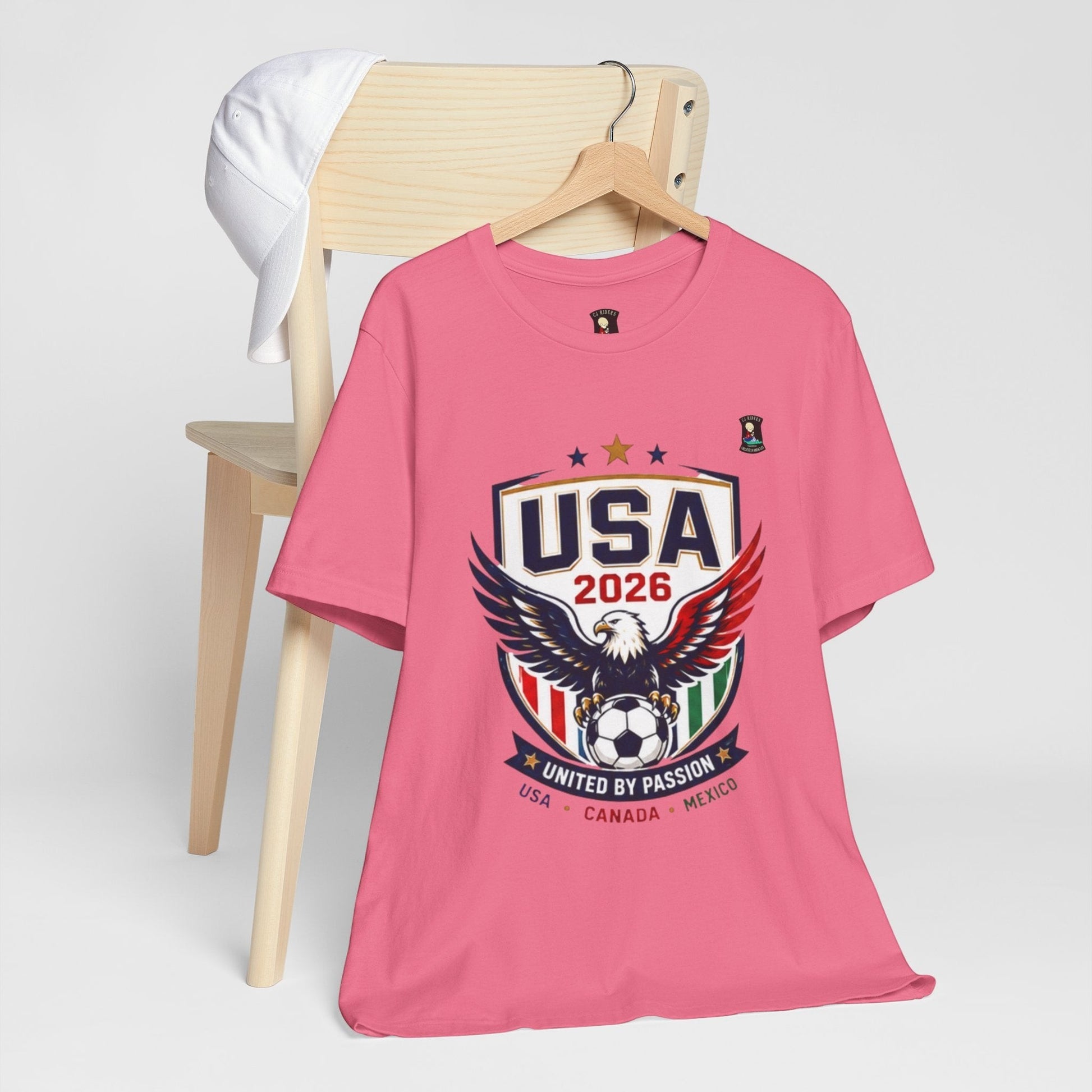 CJ Riders Foundation Store pink 100% cotton t-shirt with USA 2026 soccer shield, eagle, and "United by Passion" graphic.