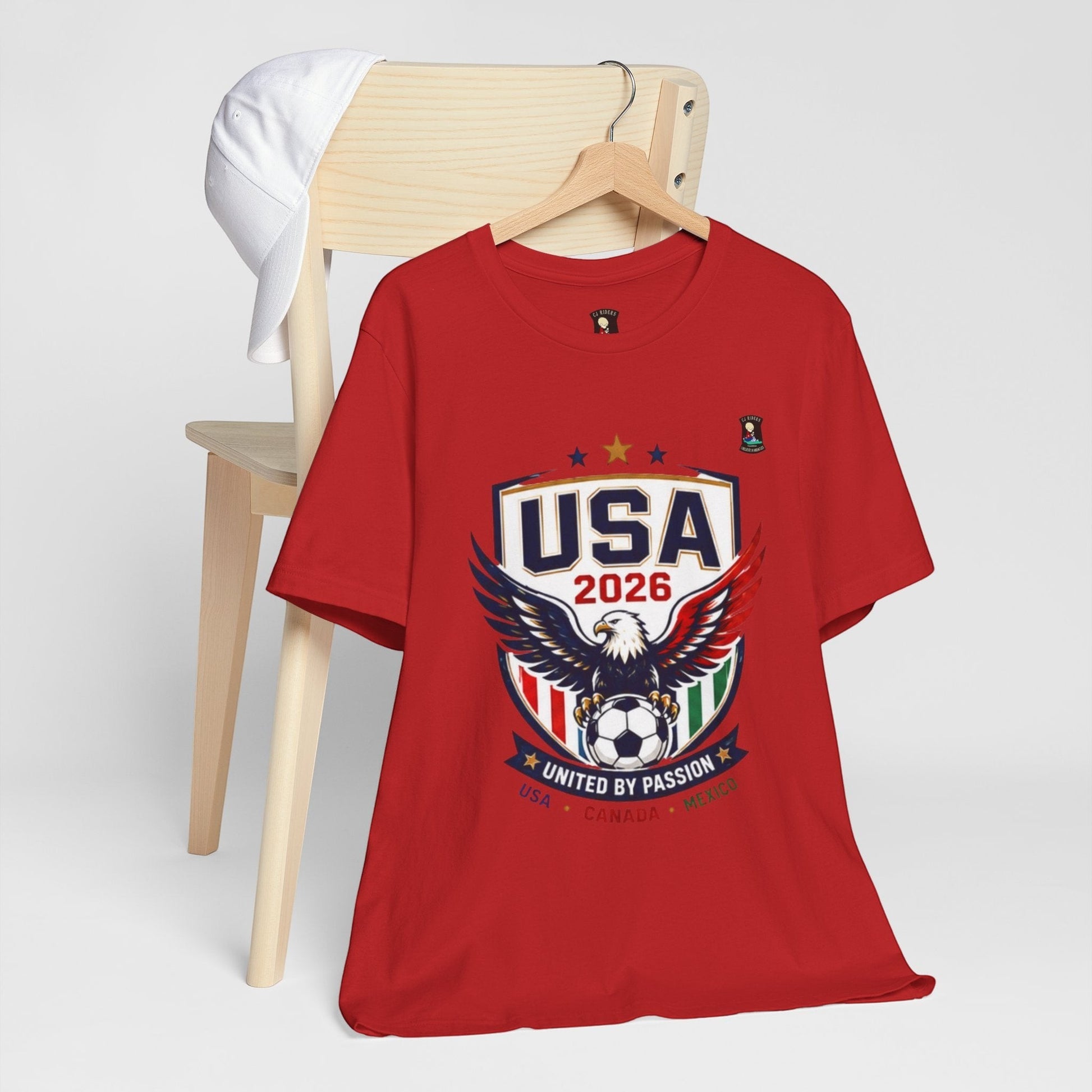 CJ Riders Foundation Store red 100% cotton t-shirt with USA 2026 eagle and soccer graphic. Features patriotic colors and "United by Passion" text.