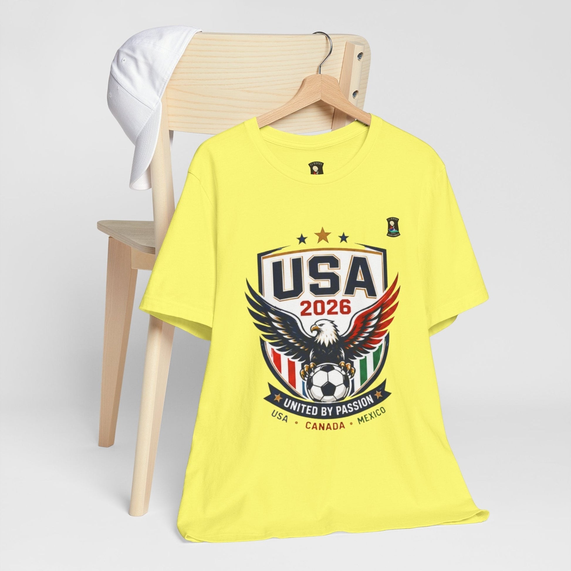 CJ Riders Foundation Store yellow 100% cotton t-shirt with USA 2026 eagle and soccer ball graphic. Features patriotic red, white, blue, and green accents.