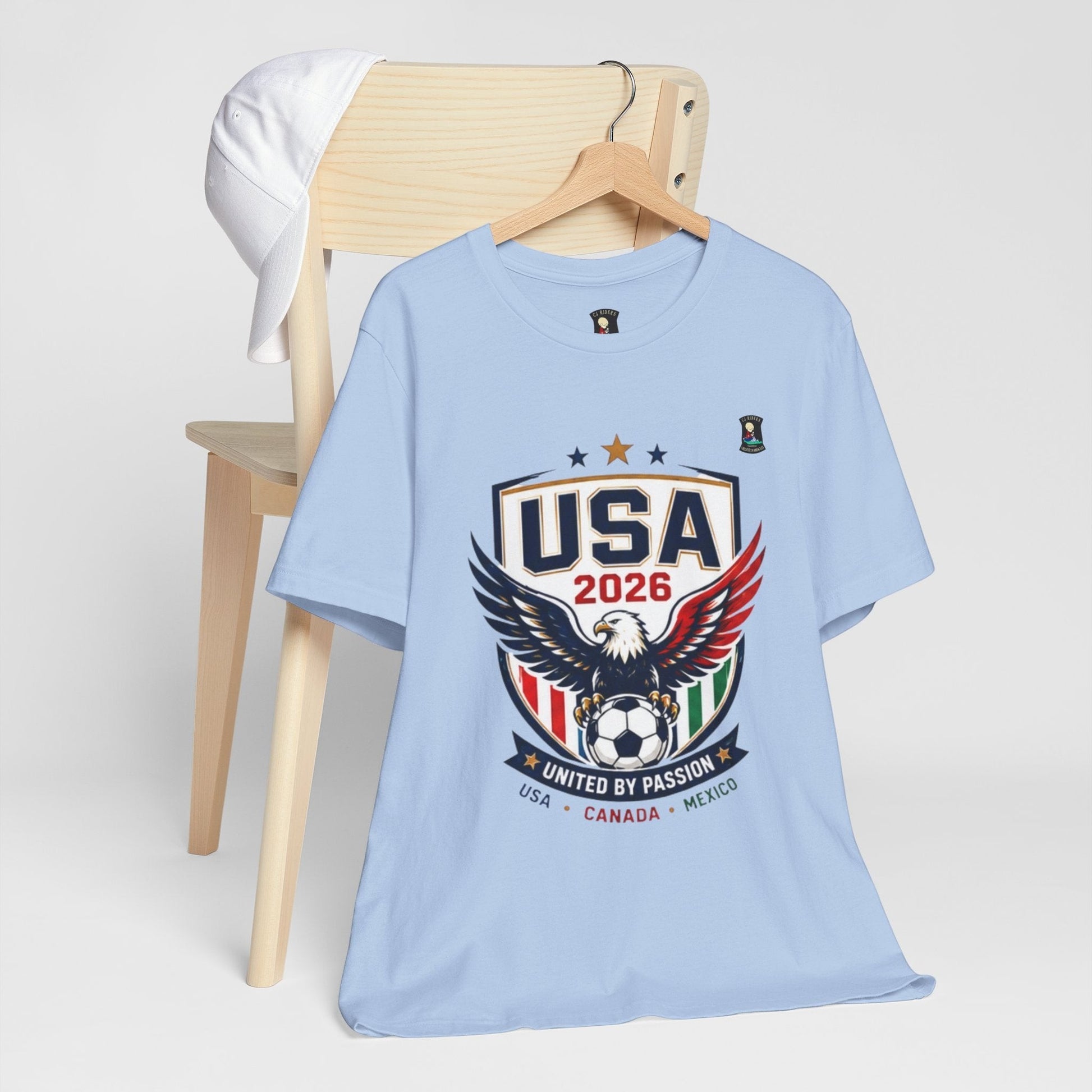 CJ Riders Foundation Store light blue 100% cotton t-shirt with USA 2026 eagle graphic and "United by Passion" text.