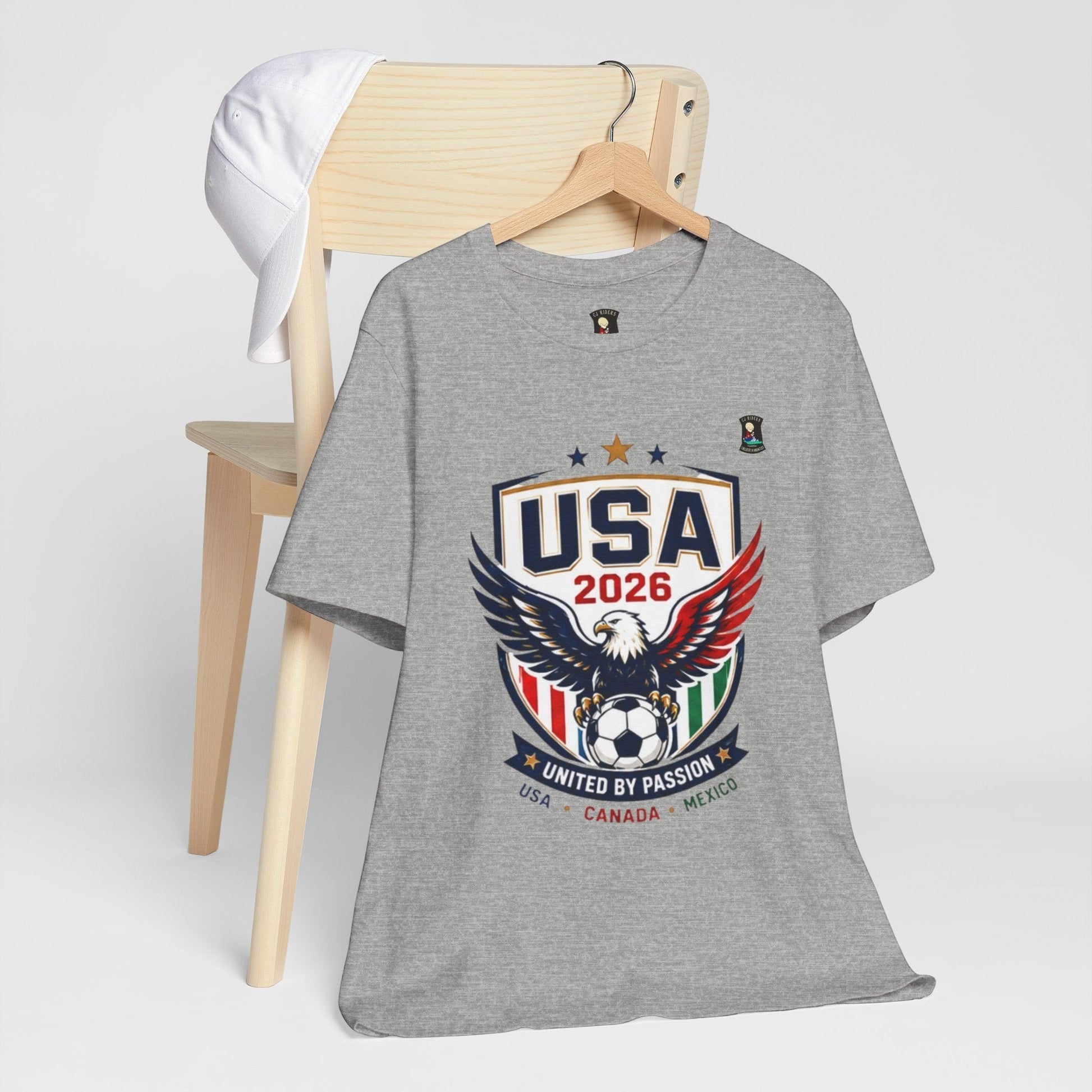 CJ Riders Foundation Store grey 100% cotton t-shirt with USA 2026 eagle and soccer ball graphic. Features patriotic red, white, blue, and green accents.