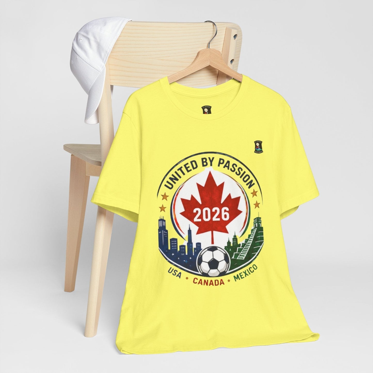 CJ Riders Foundation Store yellow 100% cotton t-shirt with a 2026 soccer crest design featuring a maple leaf, soccer ball, and city skylines.